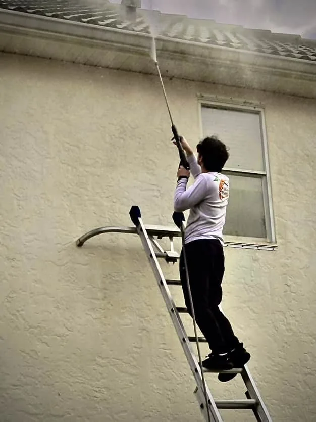 Crew member on a ladder washing a stucco wall with an extension wand before painting