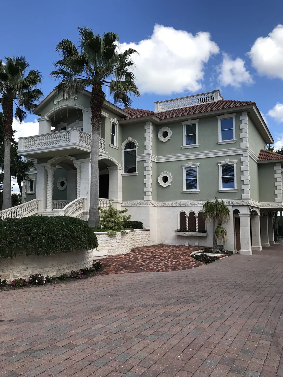 Three-story sage green stucco home with white trim above a brick paver driveway
