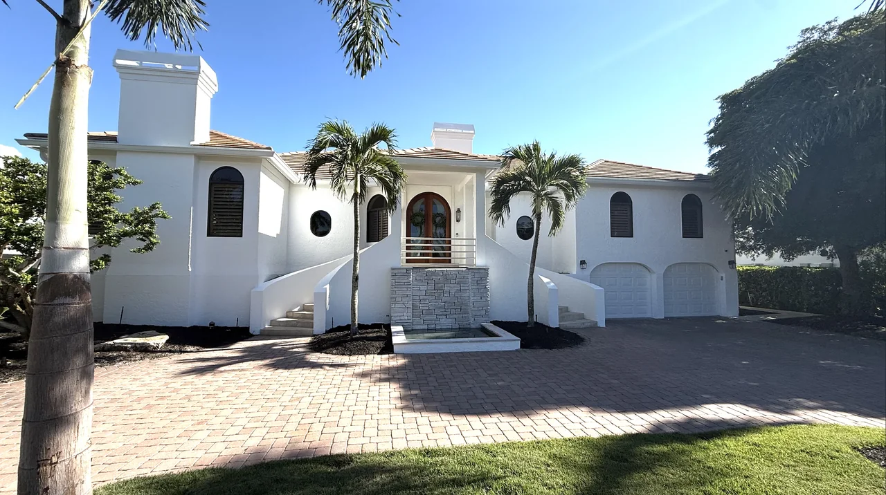 The same white stucco home seen from the paver drive, with round windows and palms